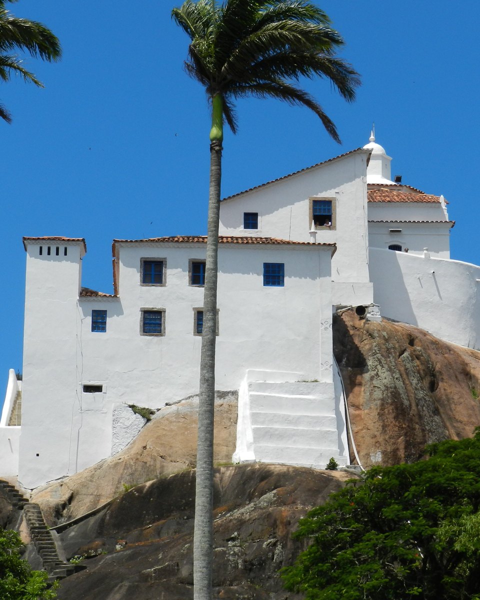 Convento da Penha, caiado de branco, sobre o rochedo, com palmeiras em primeiro plano e céu azul.