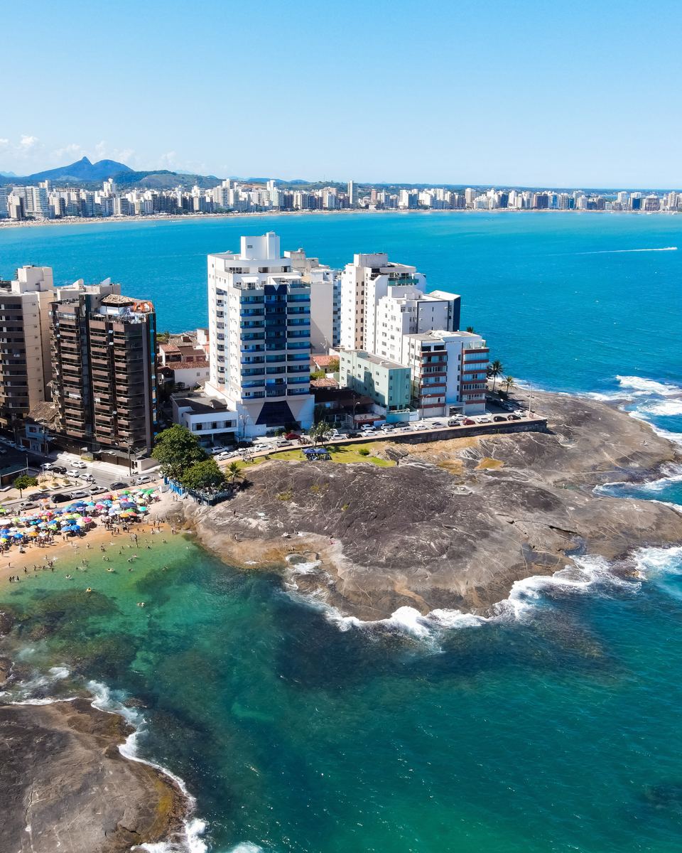 Vista aérea das praias do centro de Guarapari com banhistas, guarda-sóis coloridos e costão de pedras.
