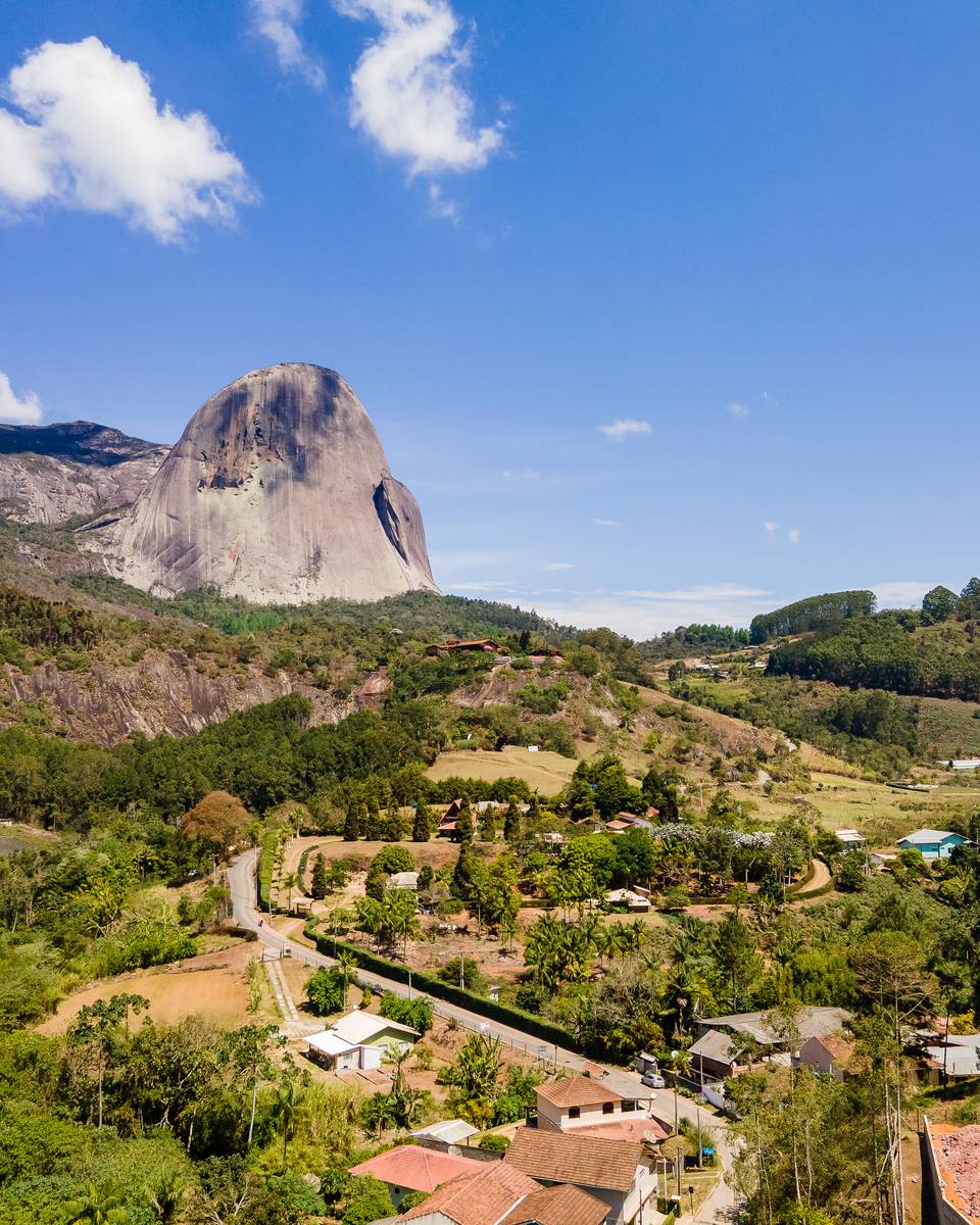 Pedra Azul erguida acima do vale, com plantações, estrada de terra e casas de telhado vermelho embaixo.