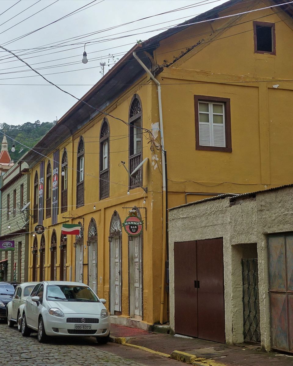 Rua do centro de Santa Teresa com casario colonial amarelo, bandeira italiana e morro de mata ao fundo.
