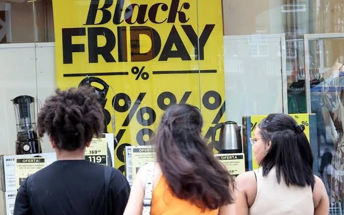 Três mulheres olhando uma vitrine com cartaz amarelo de Black Friday e produtos em oferta.