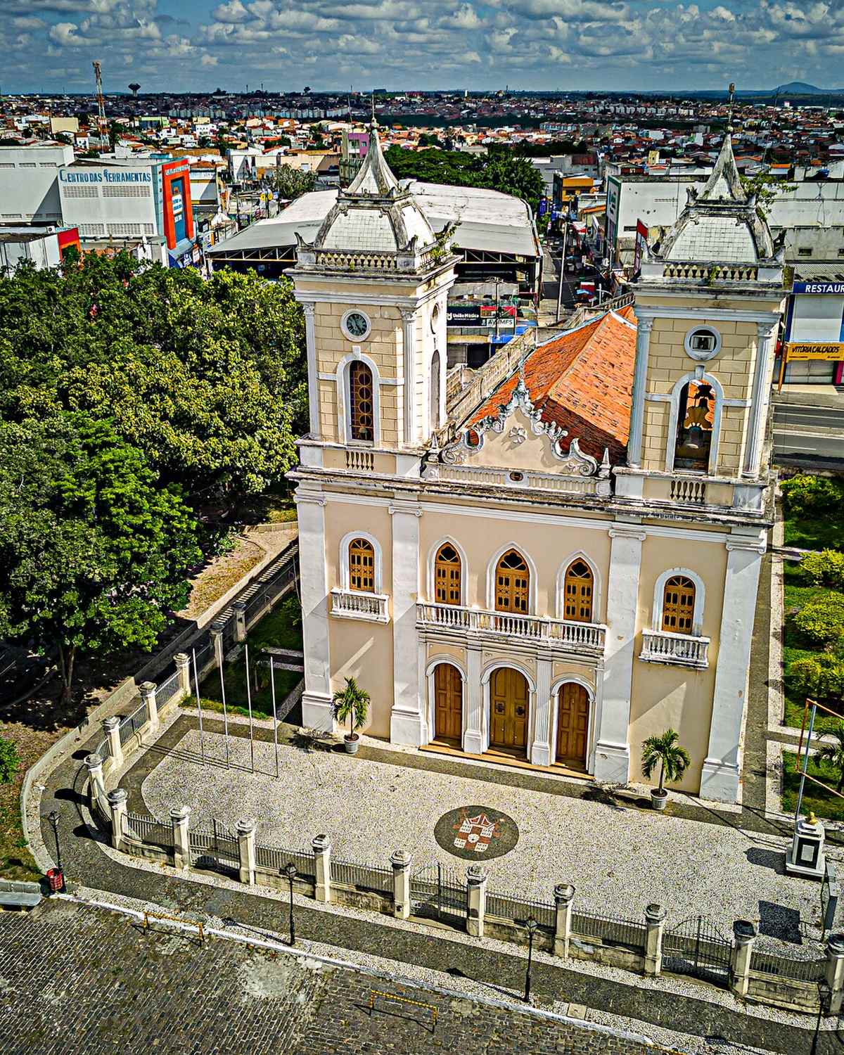 Vista aérea da Catedral Metropolitana de Senhora Sant'Ana, em Feira de Santana, de fachada clara e duas torres, cercada de árvores, com o casario e os telhados do Centro até o horizonte.