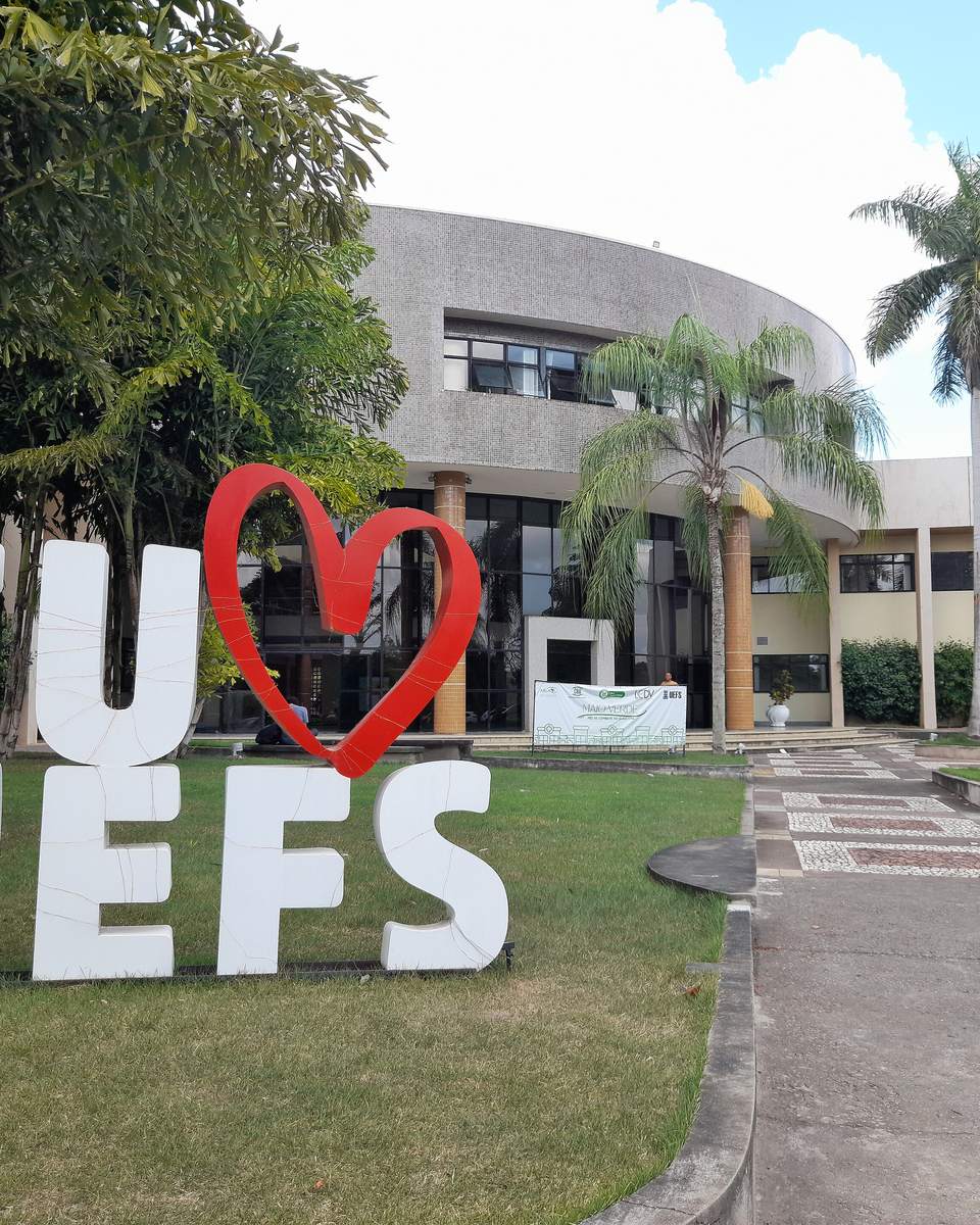 Letreiro branco com as palavras EU e UEFS e um coração vermelho no meio, no gramado em frente ao prédio da Reitoria da Universidade Estadual de Feira de Santana, com palmeiras e céu azul.