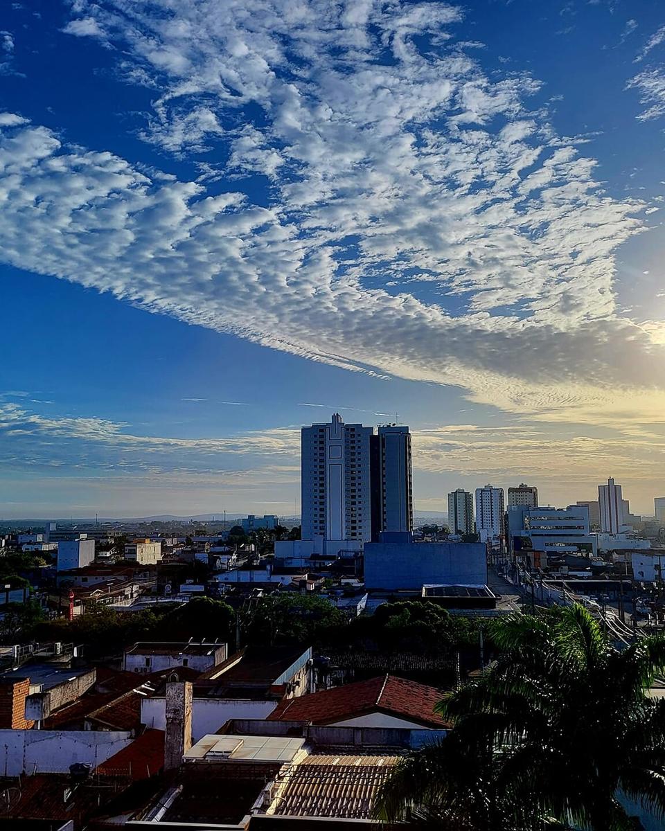 Prédios altos do bairro Santa Mônica, em Feira de Santana, vistos do alto sobre os telhados das casas, sob céu azul com nuvens em faixas e o sol no canto.