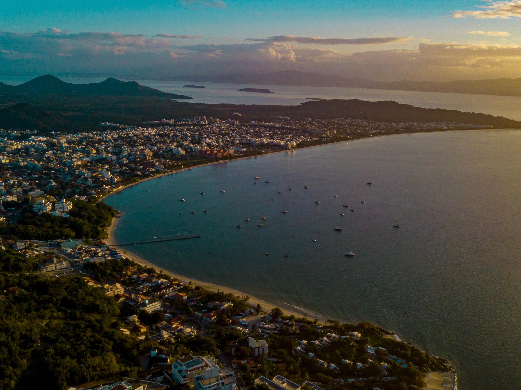Baía de Jurerê vista do alto ao entardecer, com a praia em curva, barcos ancorados na água calma, as casas entre a mata e as montanhas no horizonte sob o céu dourado.