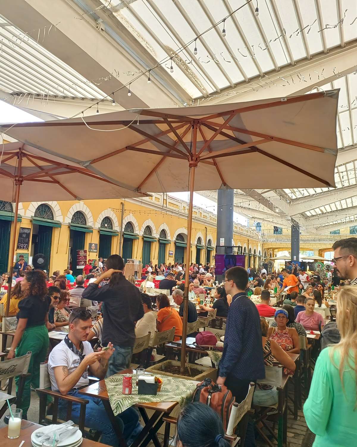Vão central do Mercado Público de Florianópolis lotado na hora do almoço, com mesas cheias sob os guarda-sóis, pessoas circulando e as arcadas amarelas do prédio ao fundo.