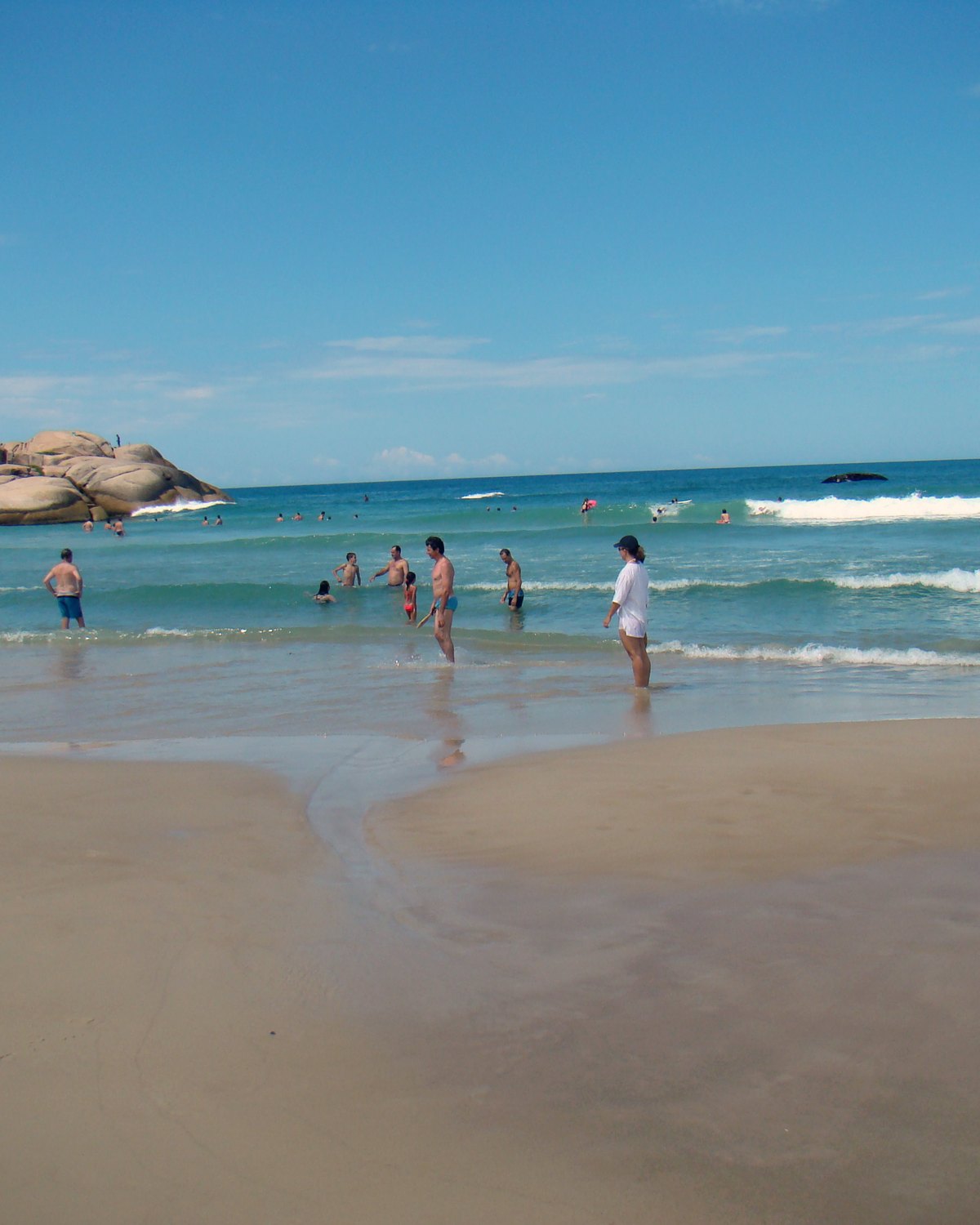 Banhistas com água pela cintura no mar da Praia da Joaquina, em Florianópolis, com a areia molhada em primeiro plano e as pedras do costão ao fundo sob céu azul.