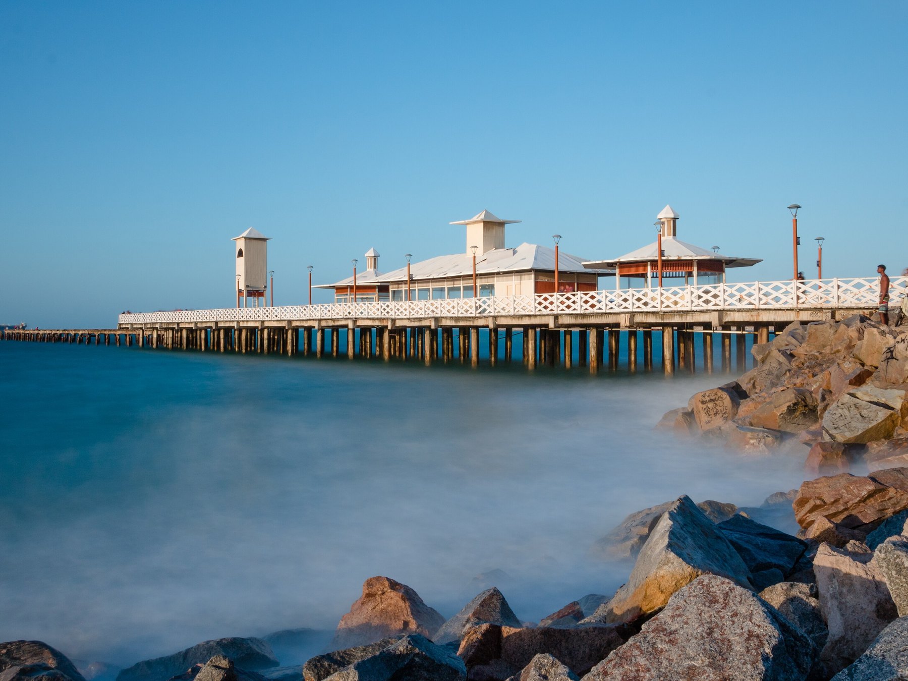 Ponte dos Ingleses avançando sobre o mar da Praia de Iracema, em Fortaleza, com os quiosques brancos de telhado vermelho, a água lisa e azul de longa exposição e as pedras escuras em primeiro plano no fim da tarde.