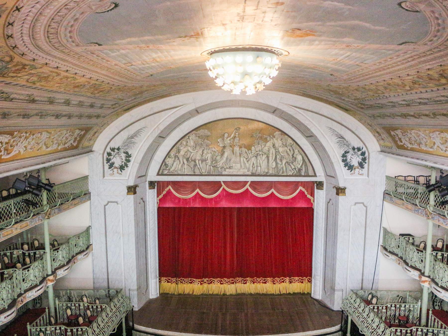 Interior do Theatro José de Alencar, em Fortaleza, visto do alto, com a boca de cena em arco, a cortina vermelha, os balcões de ferro verde em ferradura e o grande lustre de cristal pendurado no teto pintado.