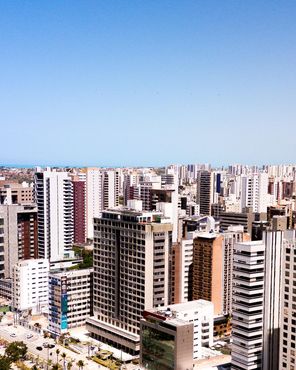 Vista aérea dos prédios altos da Aldeota e do Meireles, em Fortaleza, colados uns nos outros até o horizonte, com uma faixa de árvores e ruas em primeiro plano e o mar ao fundo sob céu azul.
