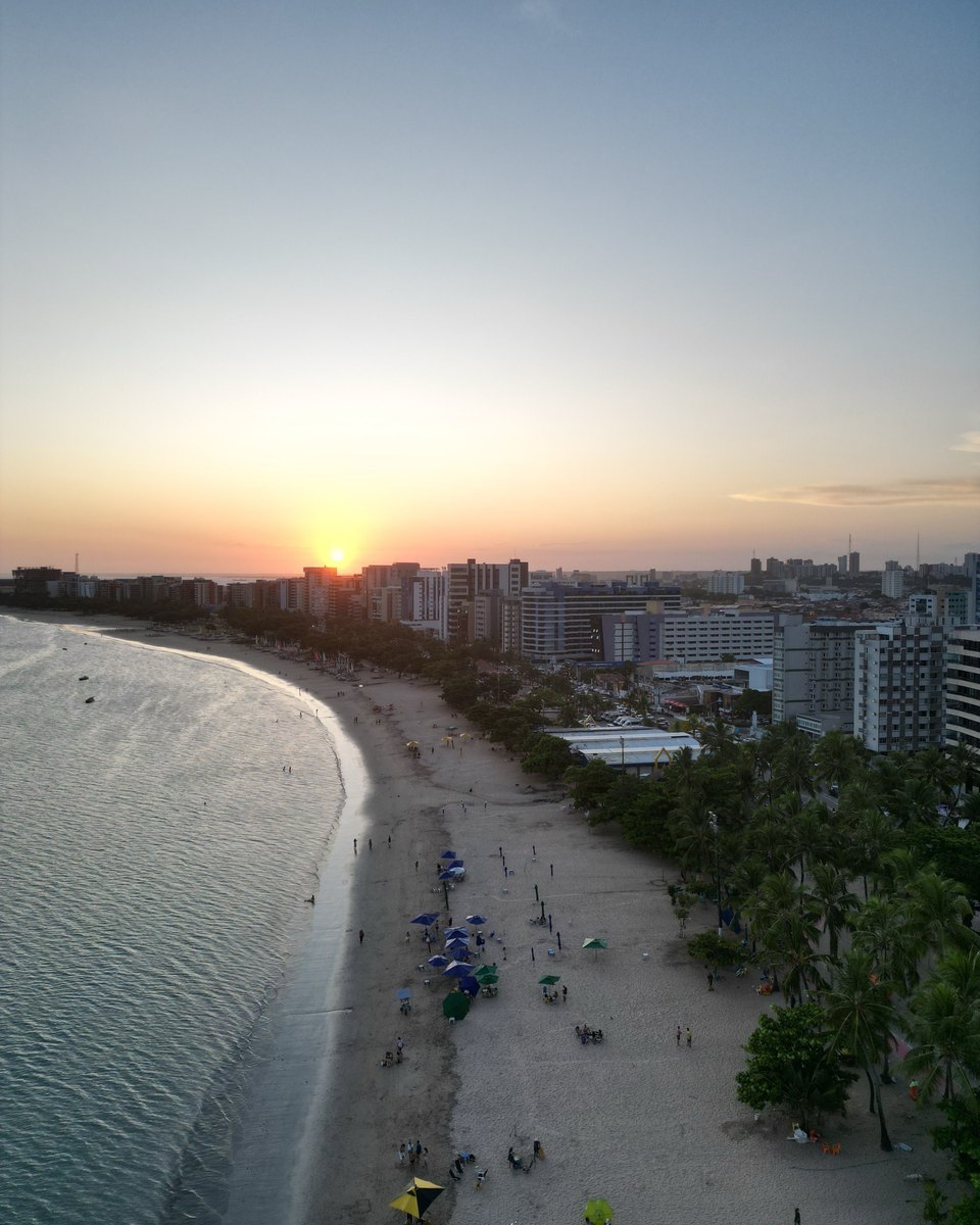 Orla de Fortaleza vista do alto ao pôr do sol, com a curva da praia, o calçadão movimentado entre as árvores e os prédios contra o céu alaranjado.