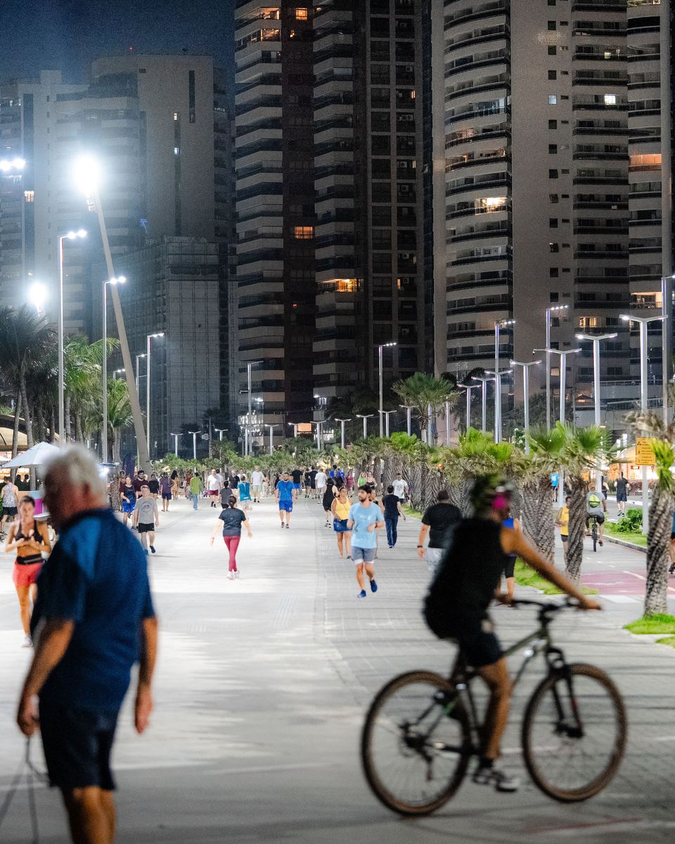 Calçadão da Avenida Beira-Mar, em Fortaleza, à noite, iluminado por postes altos, com pessoas caminhando, um ciclista passando, palmeiras e os prédios acesos ao fundo.