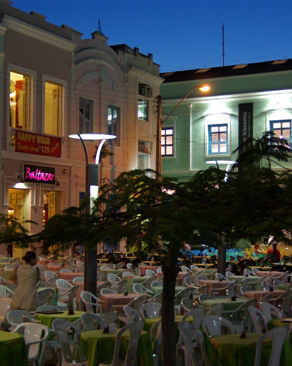 Mesas e cadeiras de bares ocupando a praça em frente aos sobrados coloridos e iluminados perto do Centro Dragão do Mar, em Fortaleza, no começo da noite.