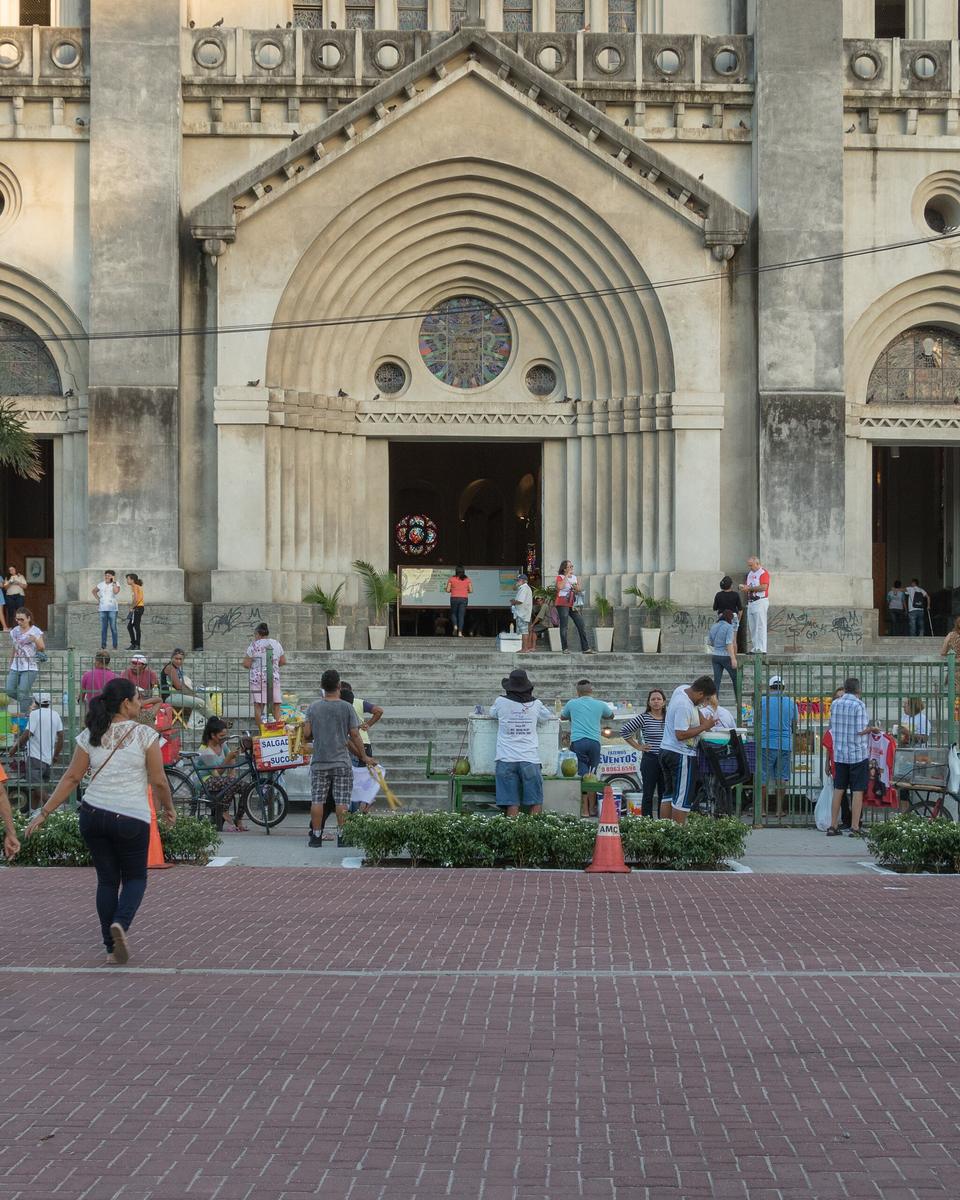 Fachada de pedra cinza da Catedral Metropolitana de Fortaleza, com portais em arco e rosácea, e pessoas atravessando a praça de piso vermelho em frente.
