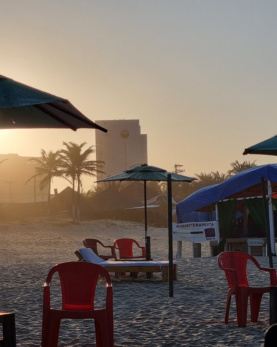 Pôr do sol numa barraca da Praia do Futuro, em Fortaleza, com guarda-sóis escuros, mesas e cadeiras vermelhas de plástico na areia e coqueiros contra o céu dourado.