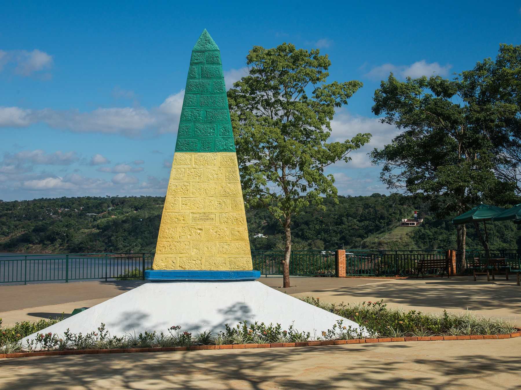 Obelisco verde e amarelo do Marco das Três Fronteiras sob o céu azul, com a praça, as árvores e o rio com a margem argentina ao fundo.