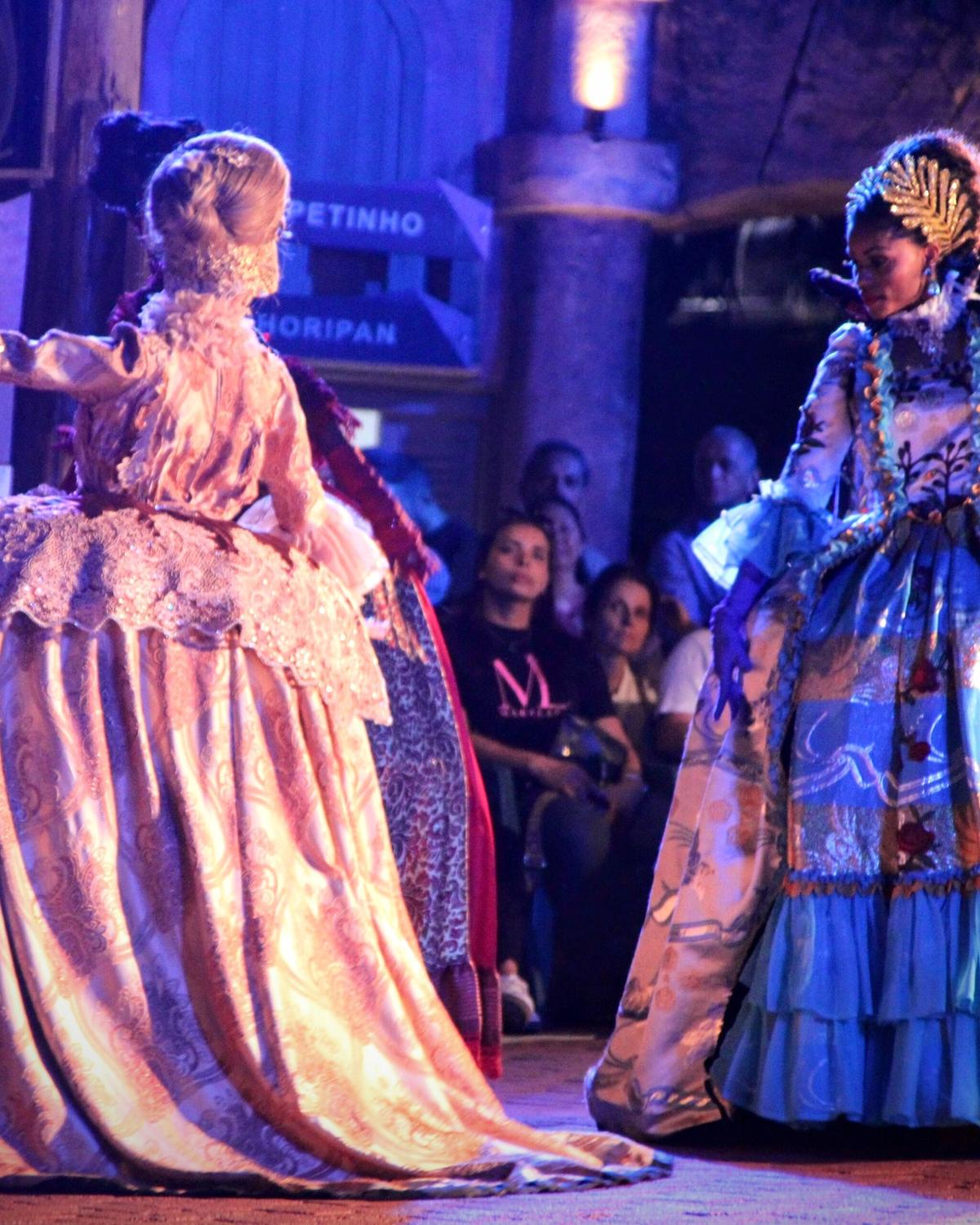Duas bailarinas com vestidos de época bordados e perucas dançando num palco iluminado de azul, com o público sentado assistindo, no Marco das Três Fronteiras.