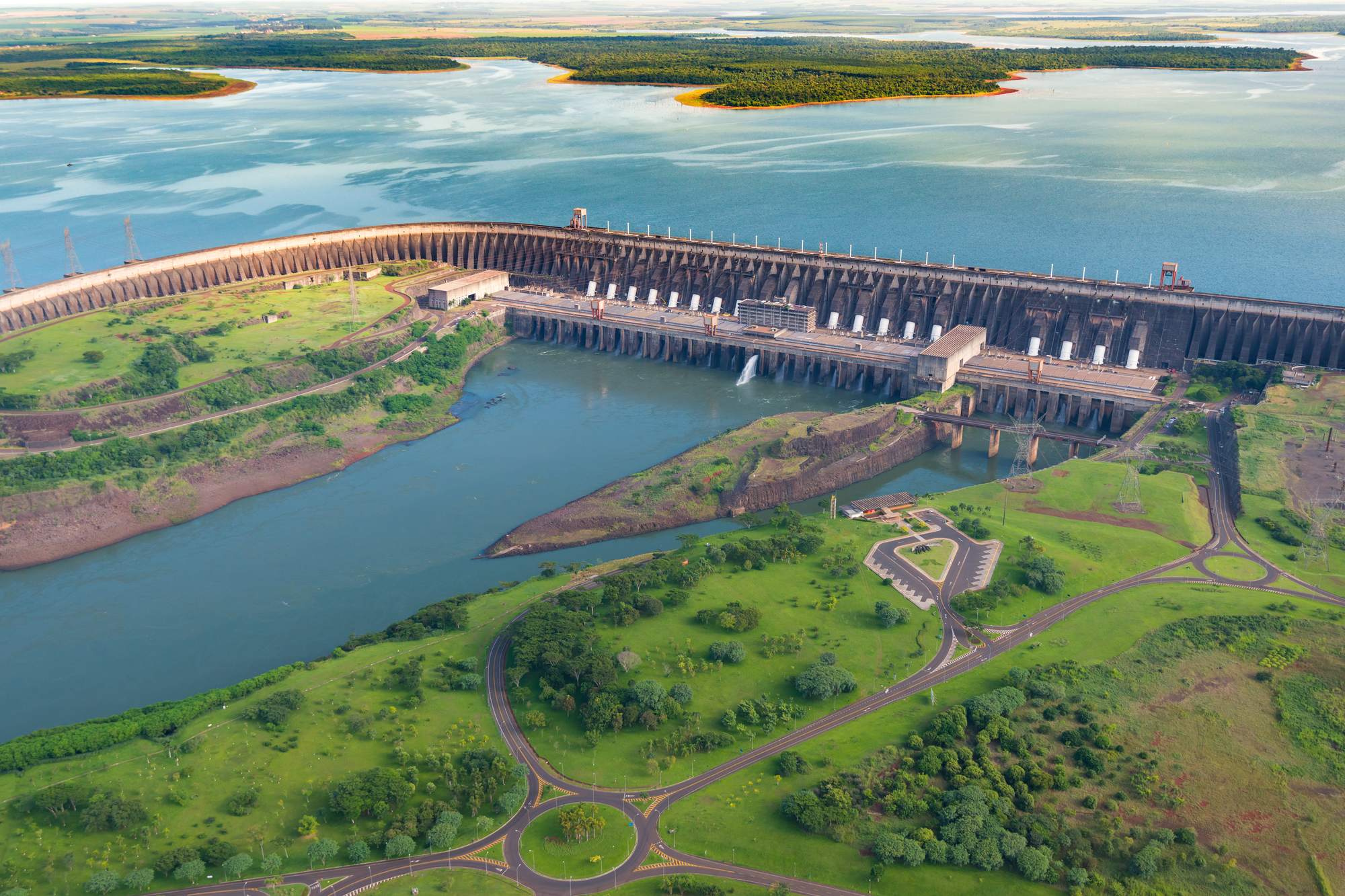 Vista aérea da usina de Itaipu, em Foz do Iguaçu, com a barragem curva segurando o lago azul, o vertedouro soltando água no rio Paraná e o gramado verde cortado por estradas em primeiro plano.
