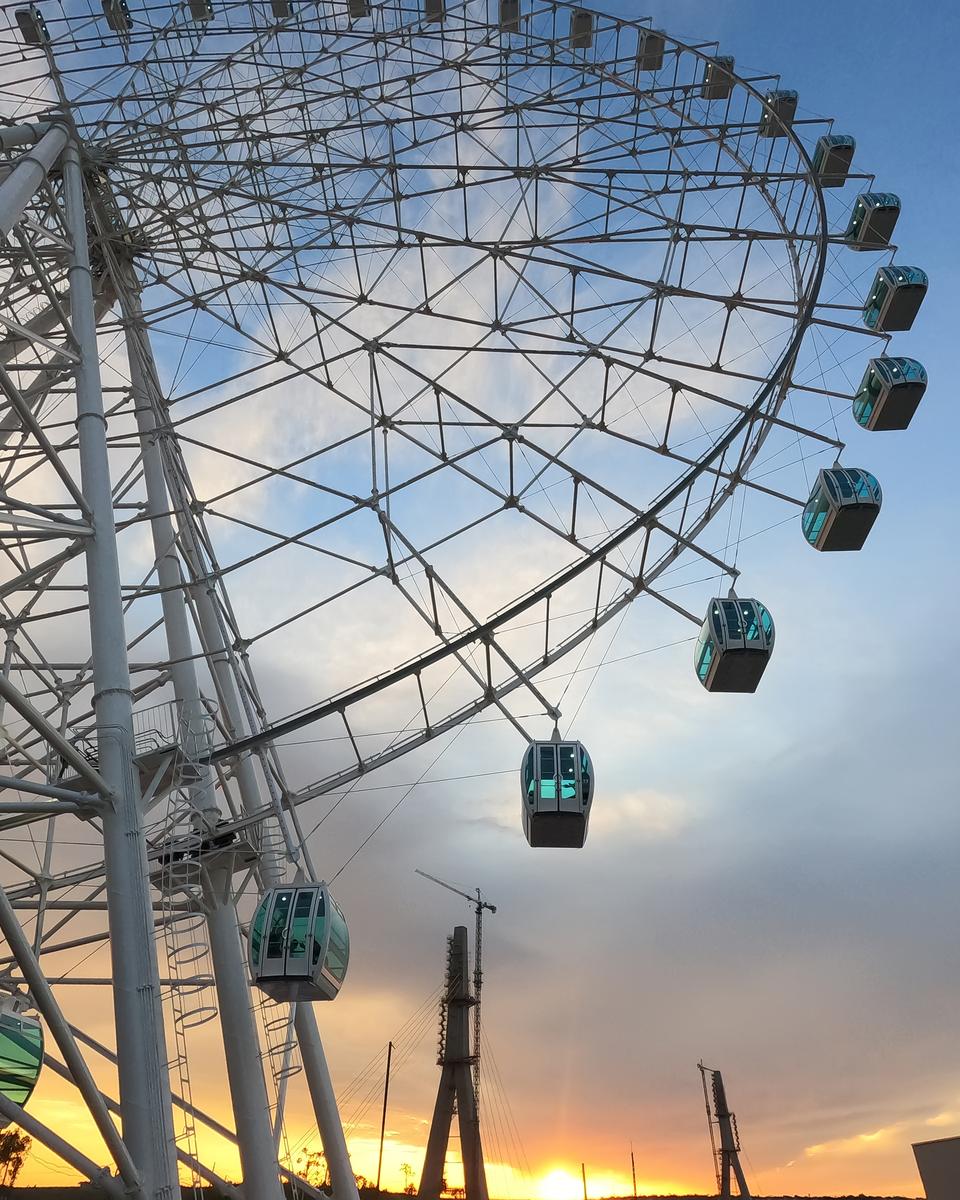 Roda-gigante branca de Foz do Iguaçu vista de baixo, com as cabines de vidro e o céu azul e alaranjado do pôr do sol ao fundo.