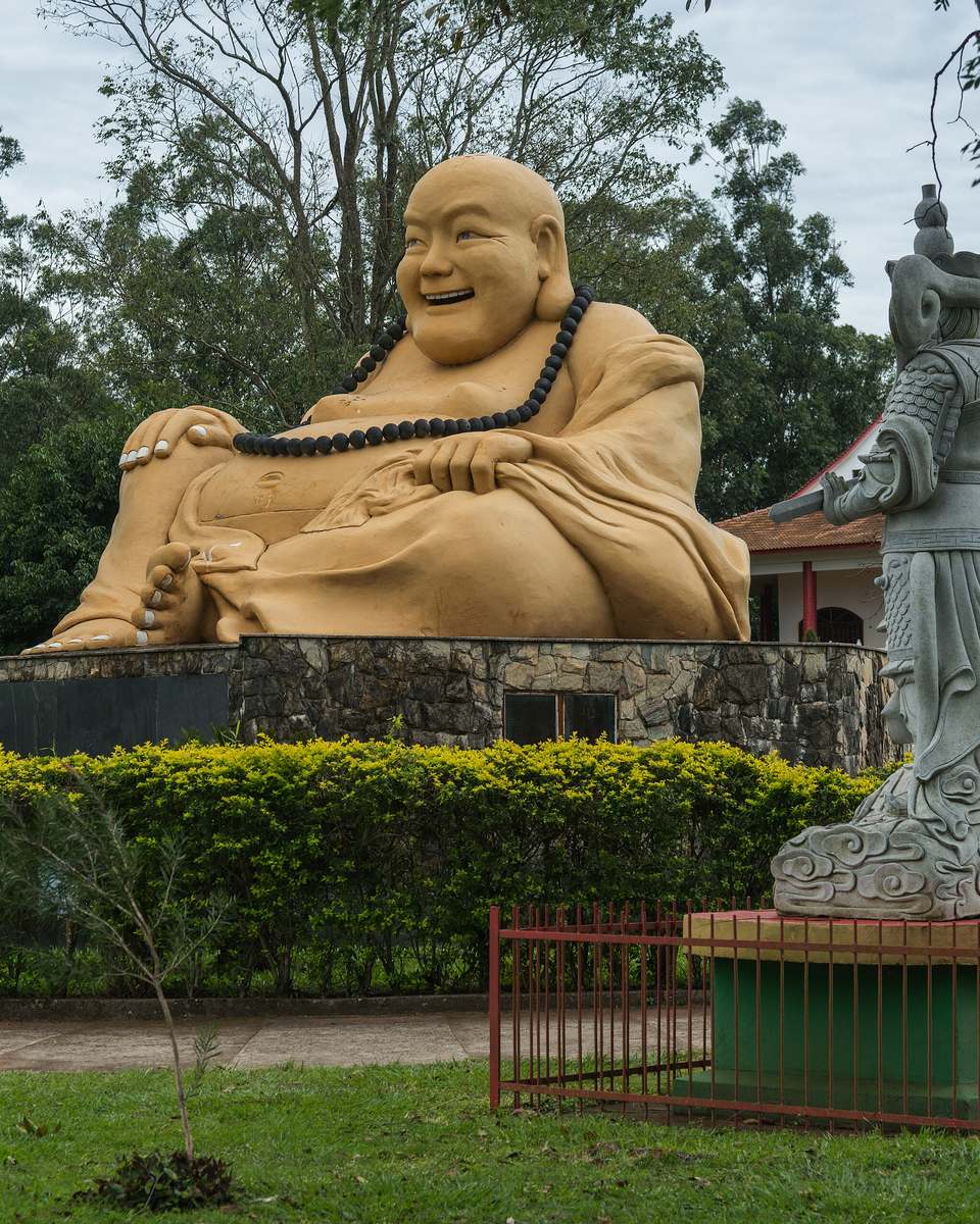 Estátua de um Buda sorridente sentado sobre um pedestal no jardim do Templo Budista Chen Tien, em Foz do Iguaçu, com uma estátua de pedra cinza ao lado, sebe verde e árvores ao fundo.