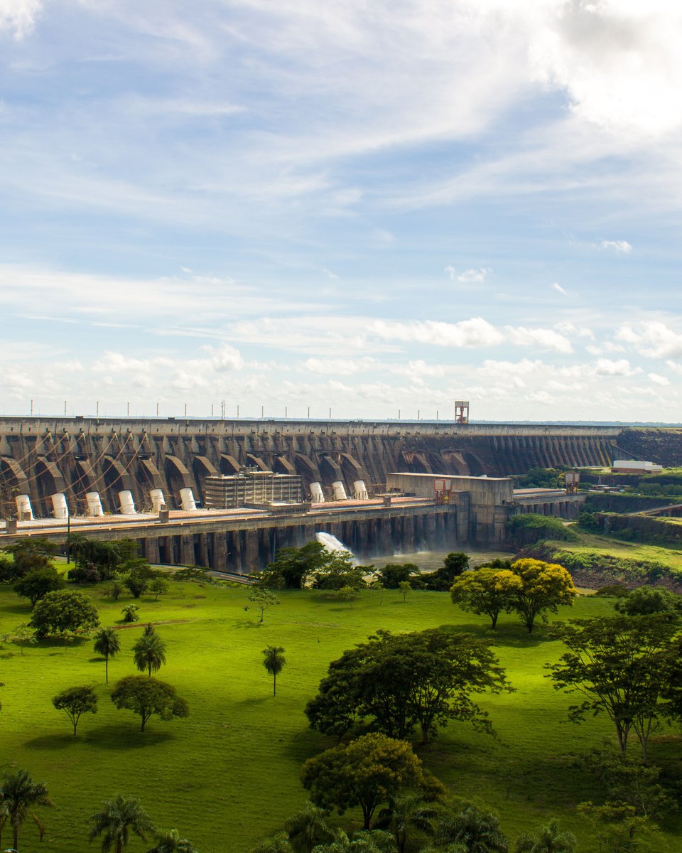 Barragem de Itaipu vista do mirante num dia de sol, com o gramado verde, palmeiras e árvores em primeiro plano e o vertedouro soltando água.