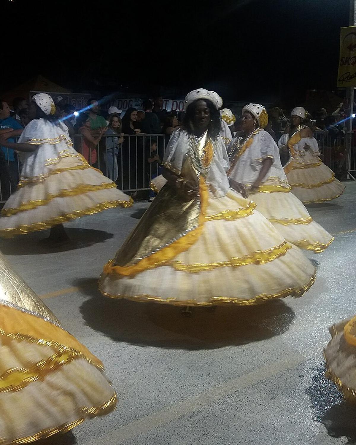 Baianas de uma escola de samba girando as saias rodadas brancas e douradas no desfile de Carnaval de Franca, à noite, com o público atrás da grade.