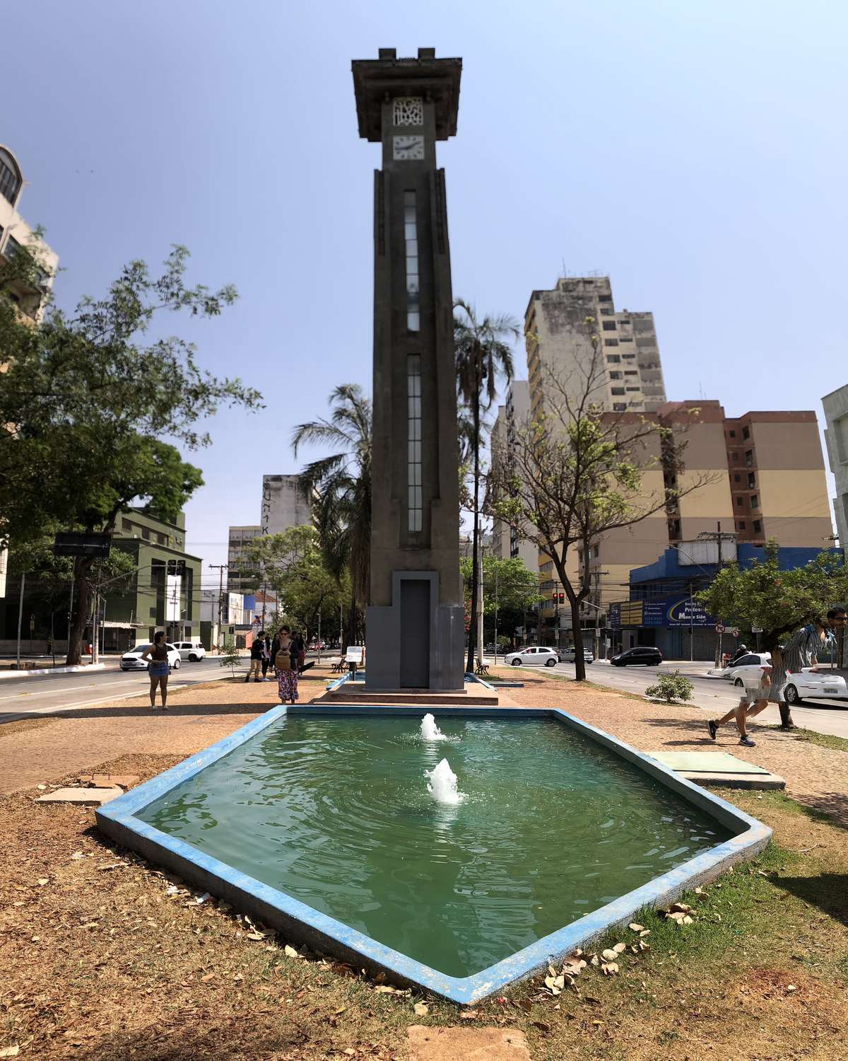 Relógio da Avenida Goiás, torre art déco de concreto no meio da praça, com o espelho d'água e a fonte na base, pessoas passando e prédios do Centro de Goiânia ao fundo, sob céu claro.