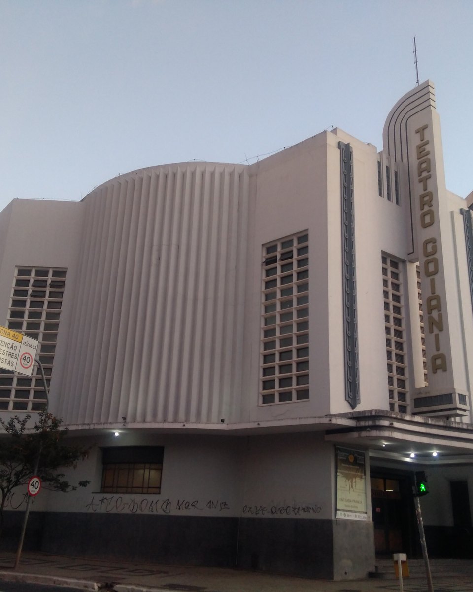 Fachada art déco do Teatro Goiânia, branca e curva, com frisos verticais, o letreiro "Teatro Goiânia" na torre da esquina e um prédio alto ao lado, num dia claro.