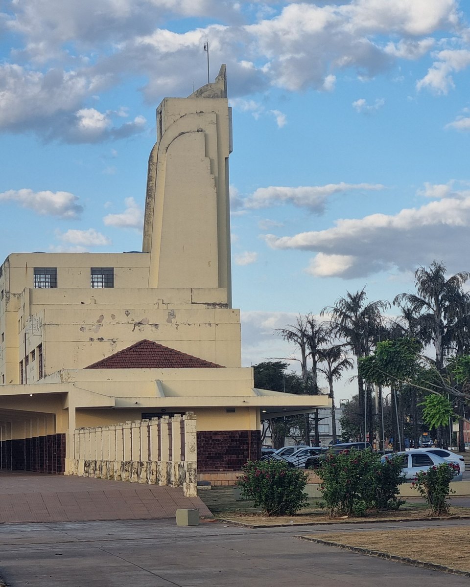 Torre art déco da antiga Estação Ferroviária de Goiânia, na Praça do Trabalhador, com a plataforma coberta, palmeiras e carros estacionados sob céu azul com nuvens.