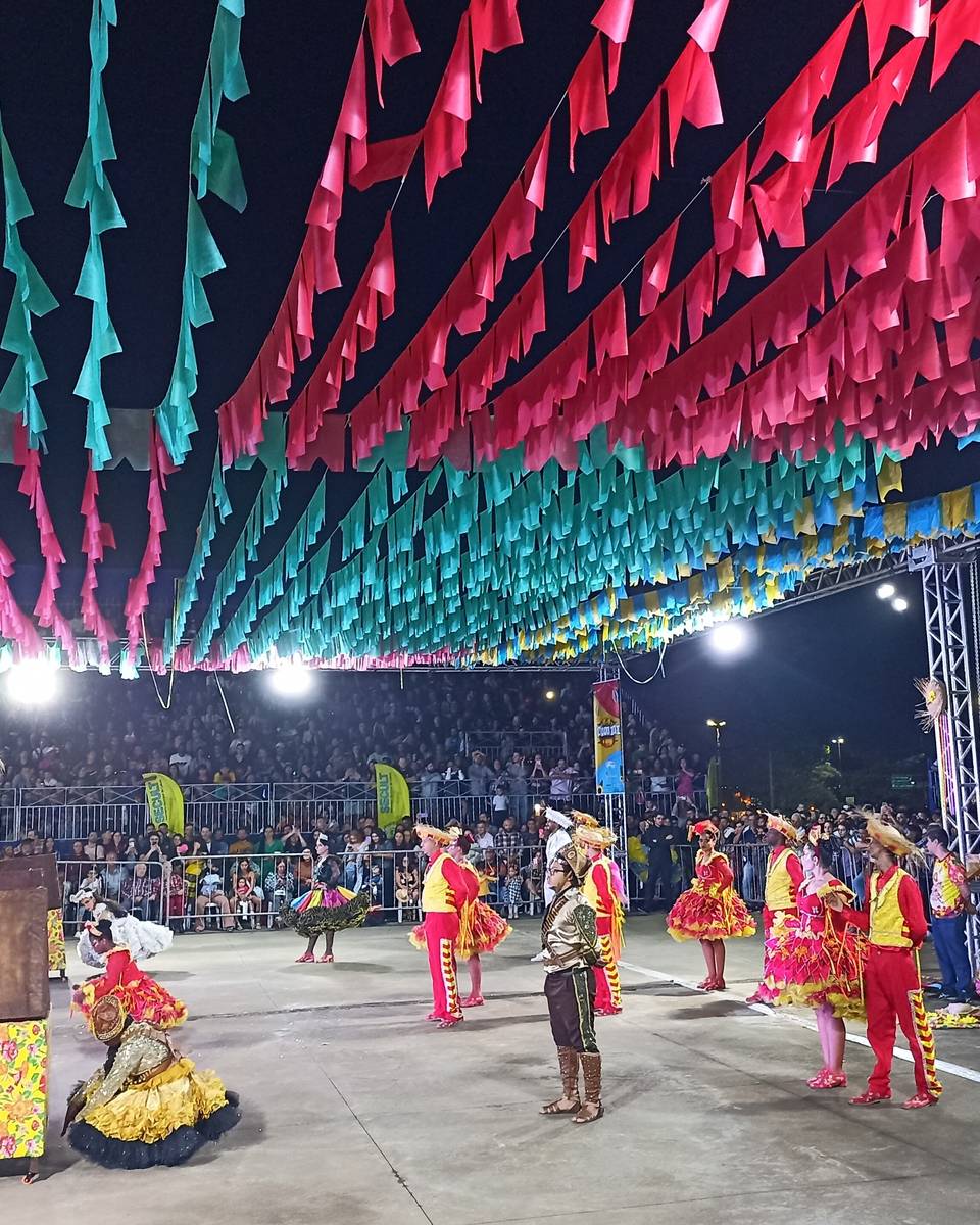 Quadrilha junina se apresentando no 20º Grande Arraiá, em Goiânia, com dançarinos de roupas amarelas e rosa, bandeirinhas rosa, azuis e amarelas cobrindo todo o teto e o público lotando as arquibancadas.