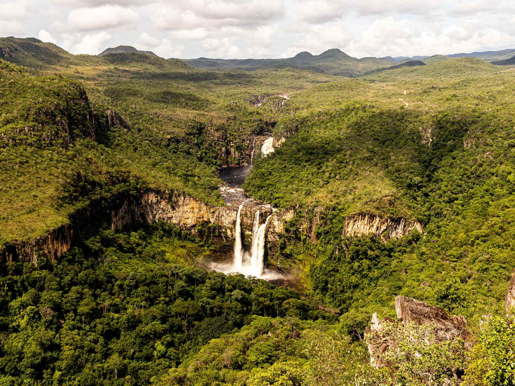 Saltos do Rio Preto vistos do alto, com duas quedas d'água caindo num cânion coberto de mata verde, paredões de pedra e as serras da Chapada dos Veadeiros no horizonte.