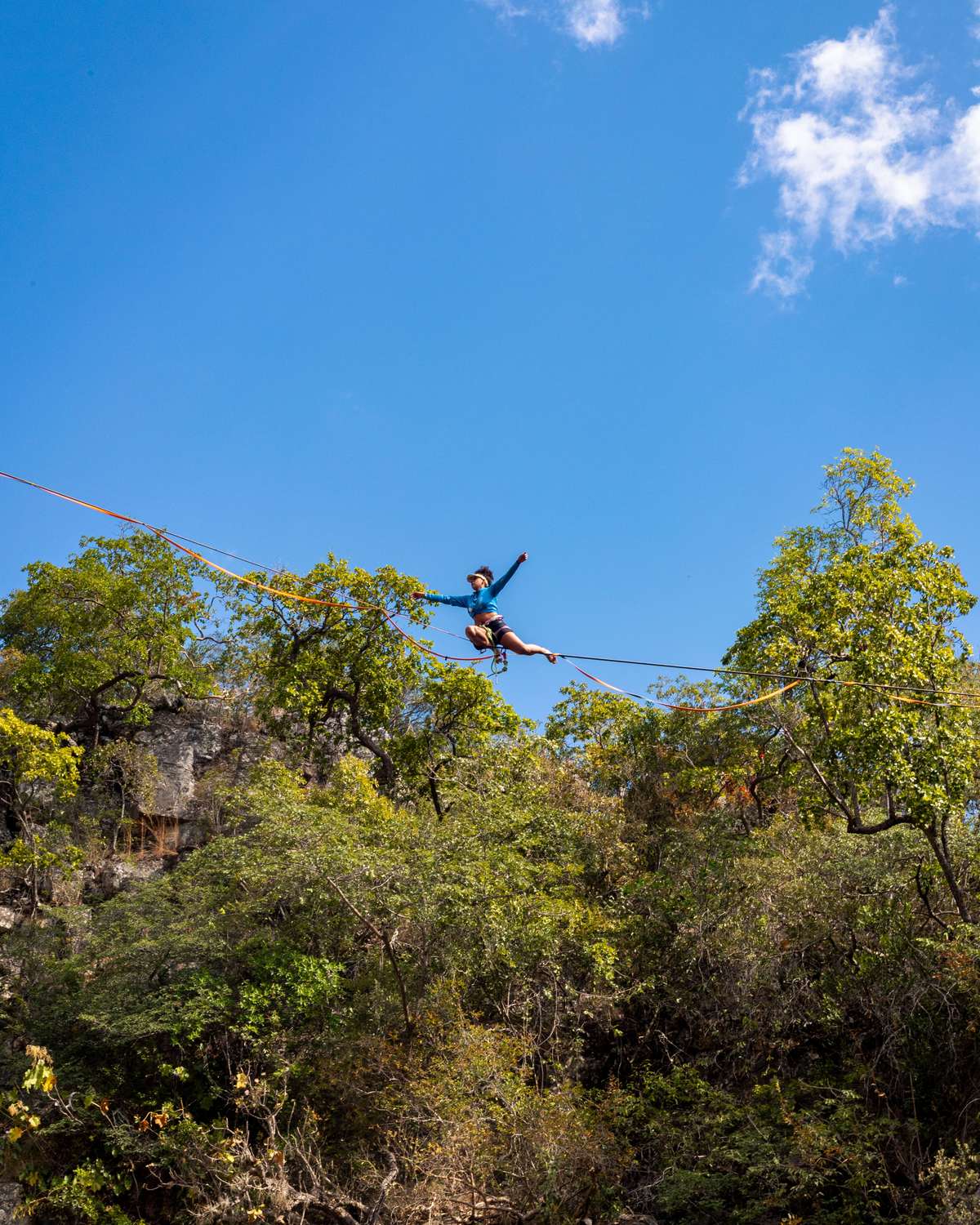 Rapaz andando numa fita de slackline esticada entre dois paredões de pedra sobre um cânion coberto de mata do cerrado, sob céu azul, em Cavalcante.