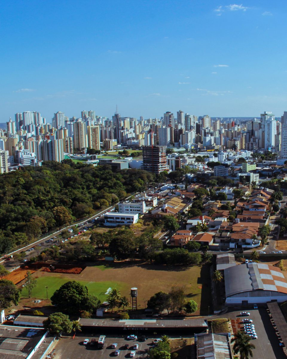 Vista aérea de Goiânia, com a fileira de prédios altos no horizonte, casas e avenidas arborizadas e uma grande mancha de mata em primeiro plano, sob céu azul.