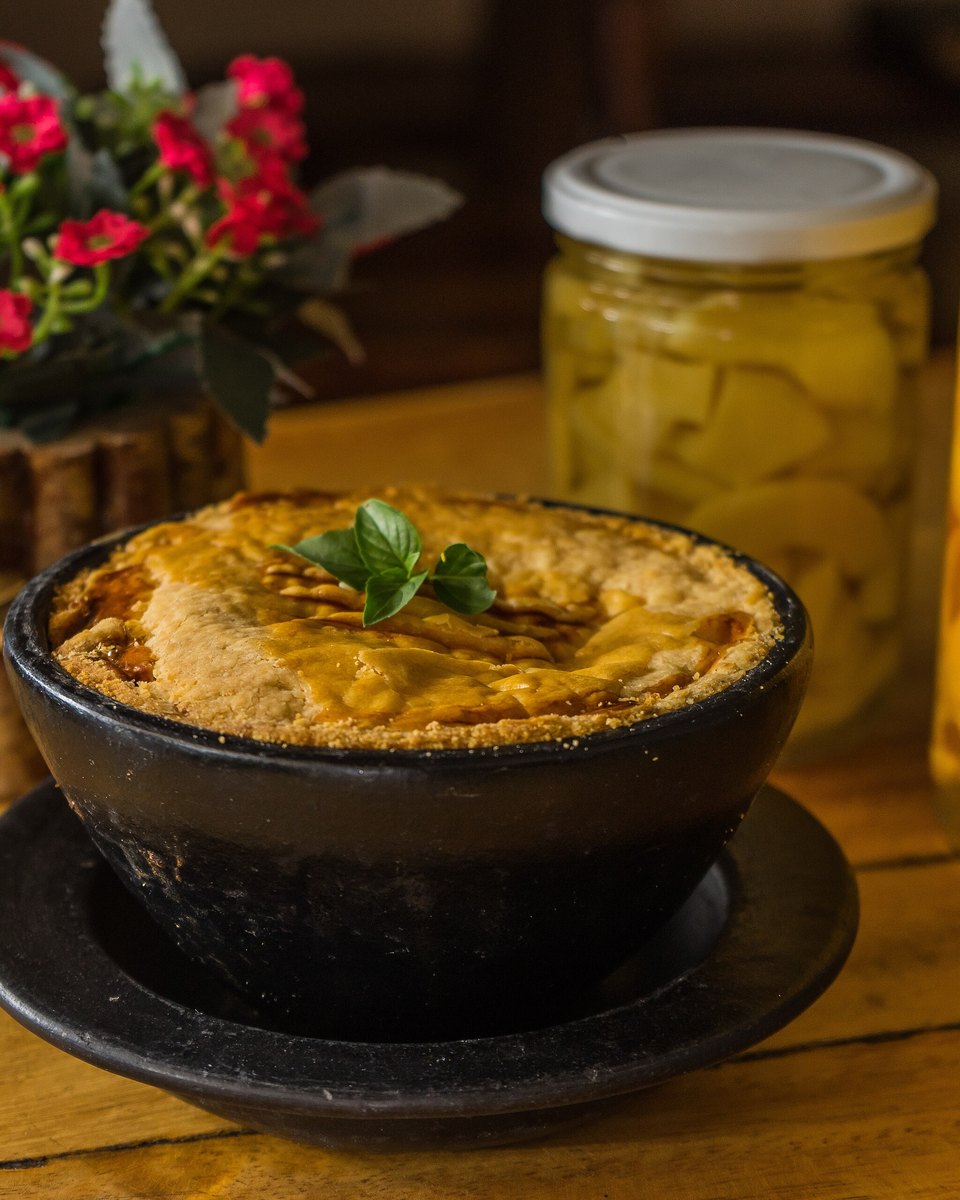 Empadão goiano servido numa cumbuca escura, com a massa dourada por cima, sobre uma mesa de madeira com potes de conserva amarela e um vaso de flores vermelhas.