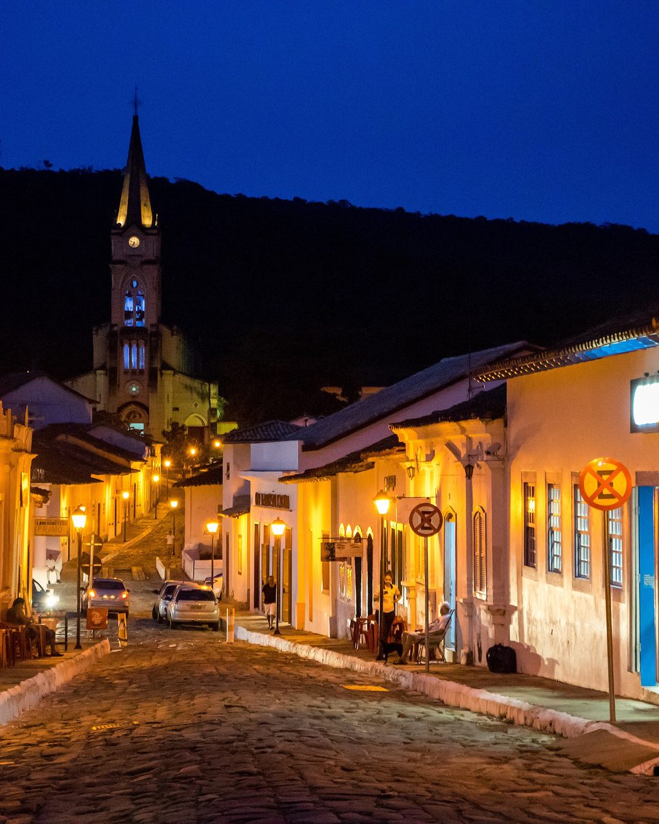 Rua de pedra do centro histórico da Cidade de Goiás ao anoitecer, com casarões coloniais iluminados, lampiões acesos e a torre de uma igreja ao fundo sob o céu azul-escuro.
