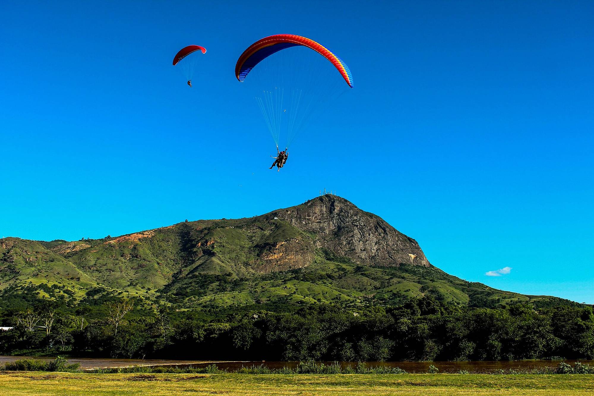 Parapente de voo duplo e outro parapente vermelho voando num céu azul sem nuvens sobre o Pico da Ibituruna, em Governador Valadares, com a mata verde e o gramado em primeiro plano.