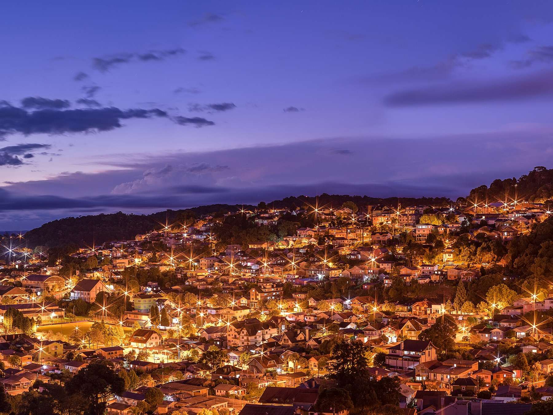Panorama de Gramado à noite, com as luzes da cidade espalhadas pelos morros e o céu azul e lilás do fim do dia.