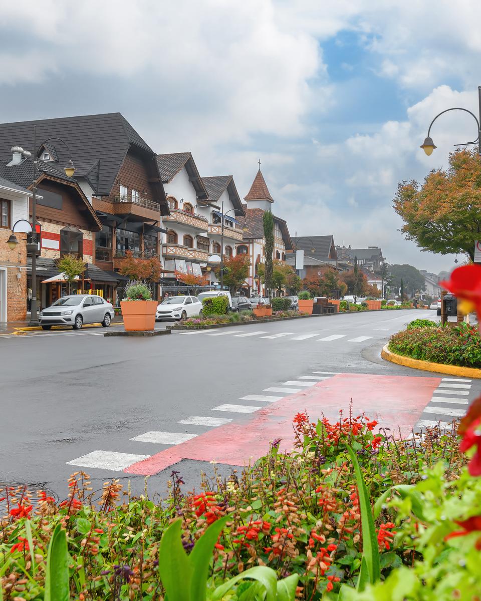 Rua do centro de Gramado num dia frio e nublado de outono, com prédios em estilo alpino, postes antigos, carros estacionados e um canteiro de flores vermelhas em primeiro plano.