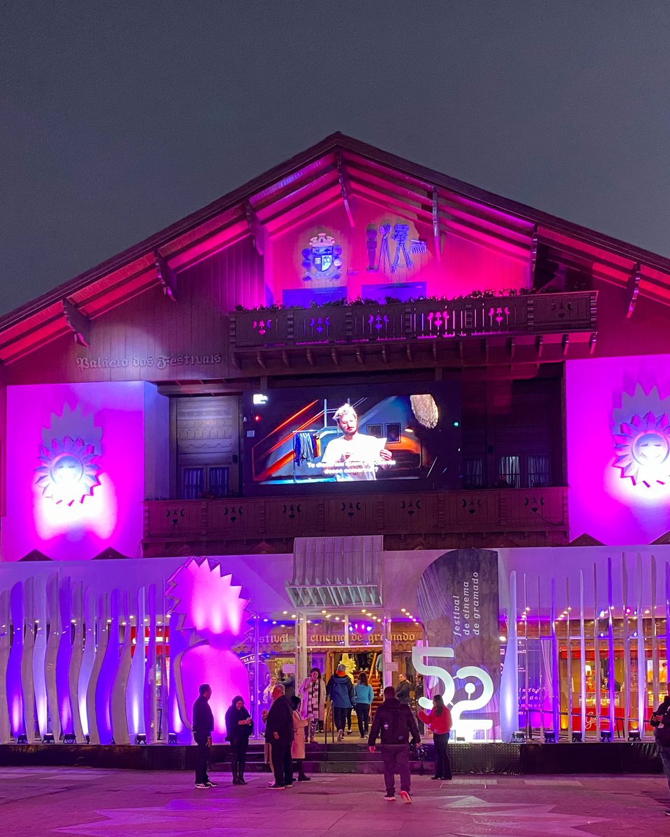 Palácio dos Festivais, em Gramado, iluminado de rosa e roxo à noite durante o Festival de Cinema, com um telão na fachada, estruturas de luz na entrada e pessoas na calçada.