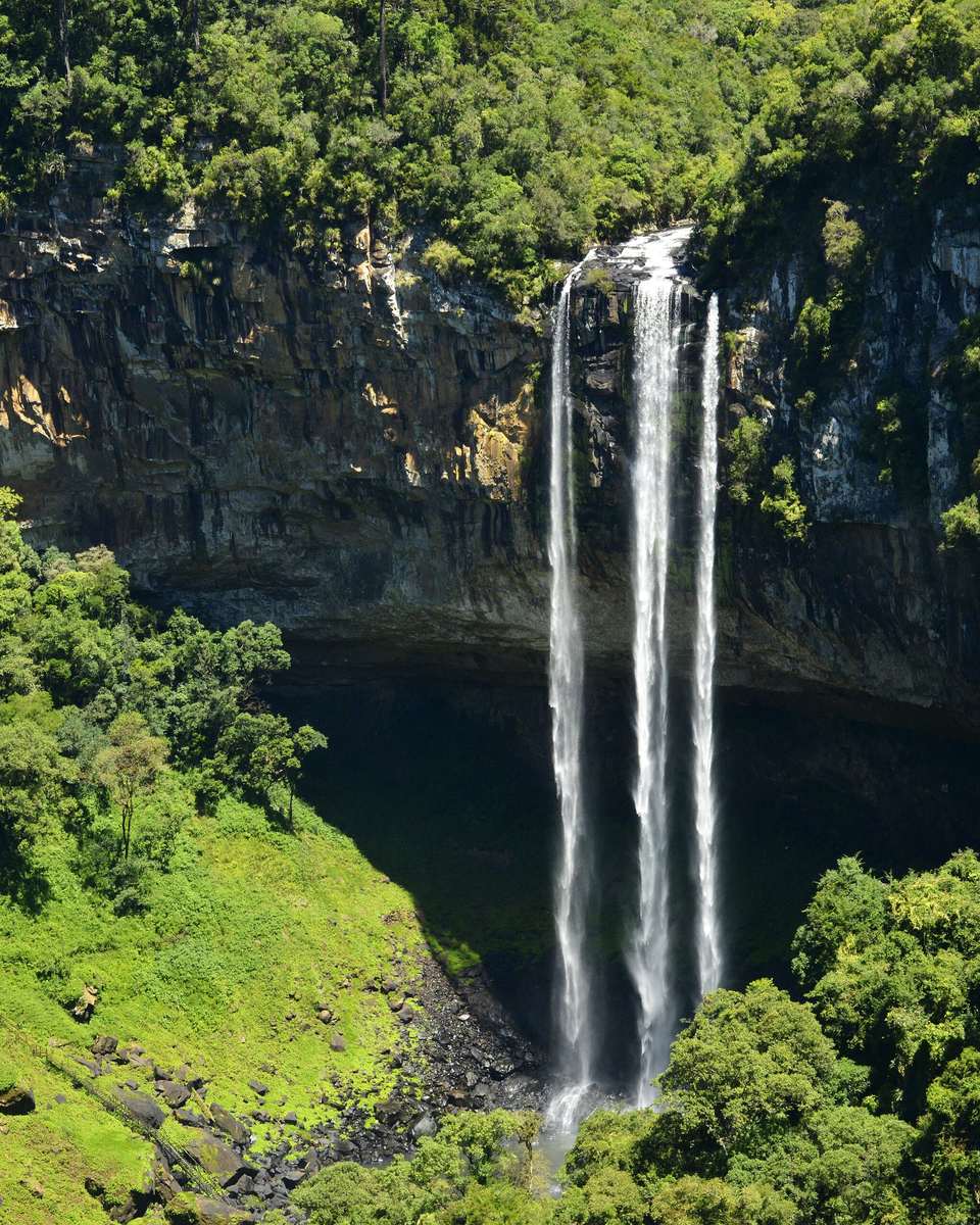Cascata do Caracol, em Canela, caindo em dois fios de água por uma parede de pedra escura cercada de mata verde, vista do alto num dia de sol.