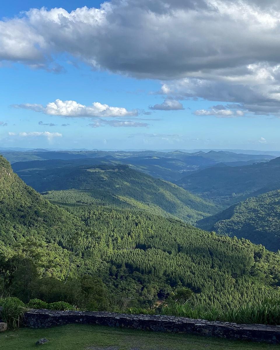 Vista do Vale do Quilombo, no interior de Gramado, com morros cobertos de mata e plantação, um paredão de pedra à esquerda e céu azul com nuvens.