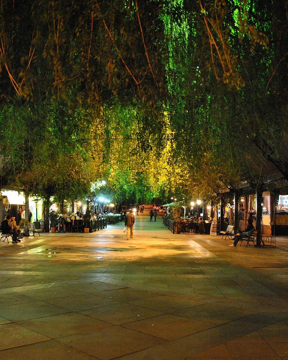 Rua Coberta de Gramado à noite, com plantas penduradas iluminadas de verde e amarelo sob o teto, calçamento de pedra, mesas de restaurante e pessoas passeando.