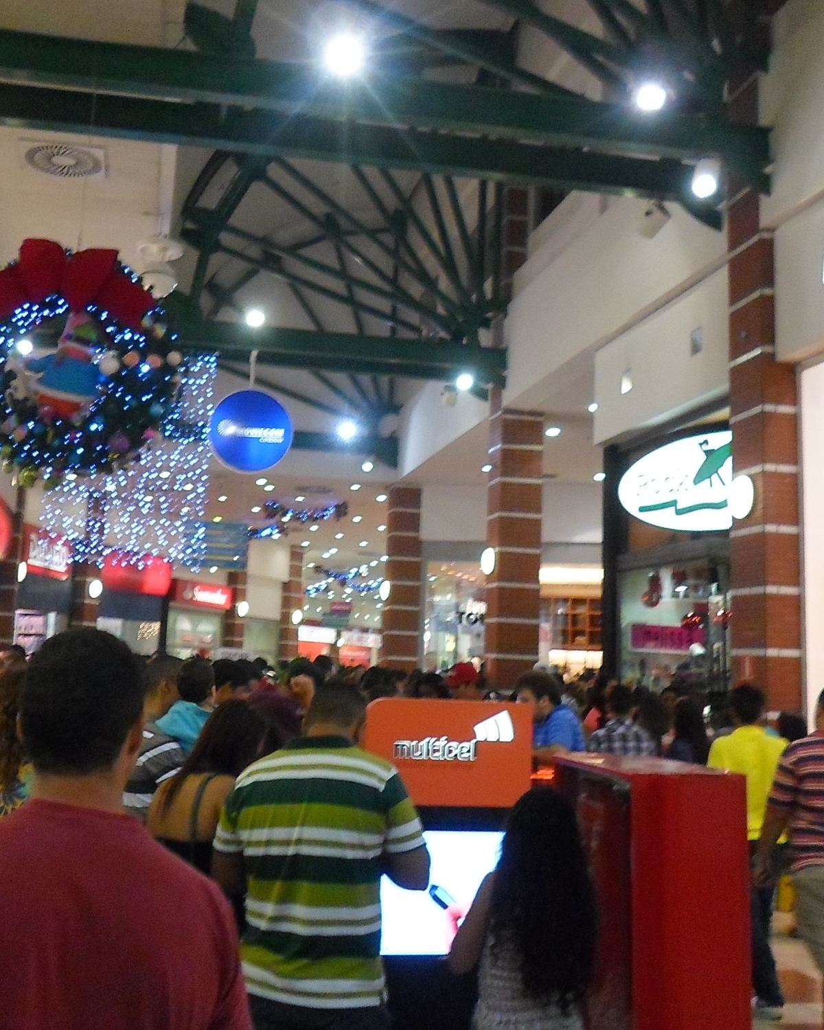 Corredor lotado do Shopping Vale do Aço, em Ipatinga, decorado para o Natal, com uma guirlanda iluminada pendurada no teto, vitrines acesas e pessoas passeando entre as lojas.