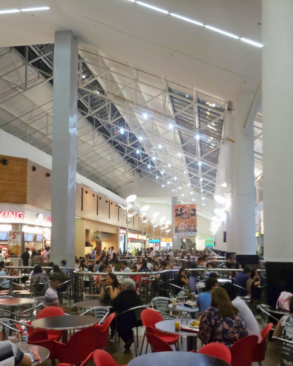 Praça de alimentação do Shopping Vale do Aço, em Ipatinga, cheia de gente sentada em mesas e cadeiras vermelhas, sob um teto alto de vidro e estrutura metálica, com balcões de lanchonetes ao fundo.