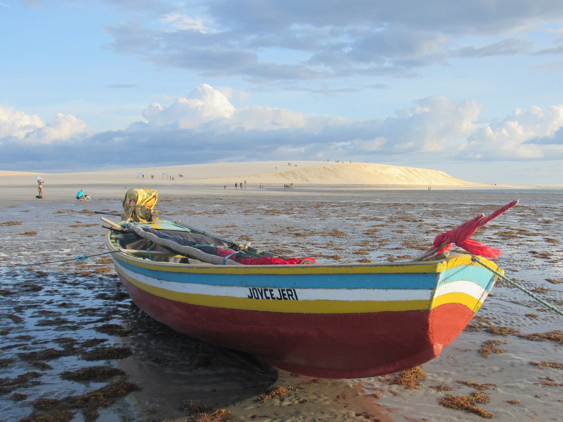 Jangada colorida de listras vermelhas, amarelas e azuis encalhada na maré baixa da praia de Jericoacoara, com poças de água no chão de areia e a Duna do Pôr do Sol ao fundo sob céu de nuvens.