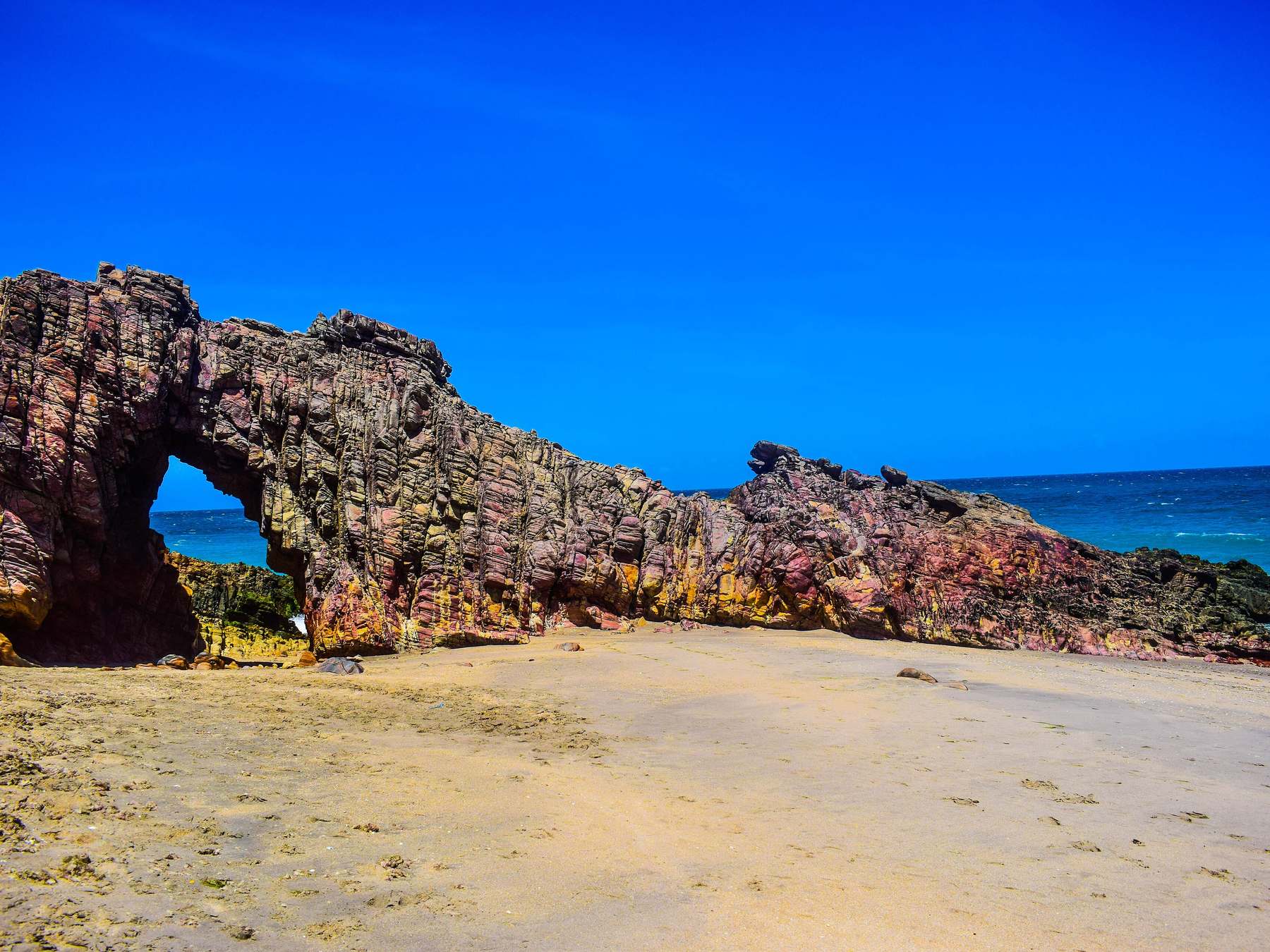 Pedra Furada de Jericoacoara, um arco de rocha em tons de rosa, roxo e amarelo sobre a areia, com o mar azul aparecendo pela abertura e o céu azul sem nuvens.