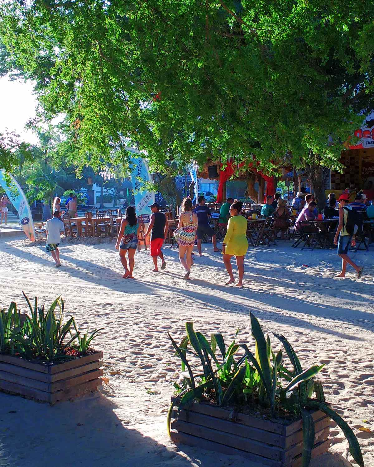 Turistas de roupa de praia caminhando pela areia clara da vila de Jericoacoara, à sombra de árvores, com vasos de plantas e restaurantes coloridos ao fundo.
