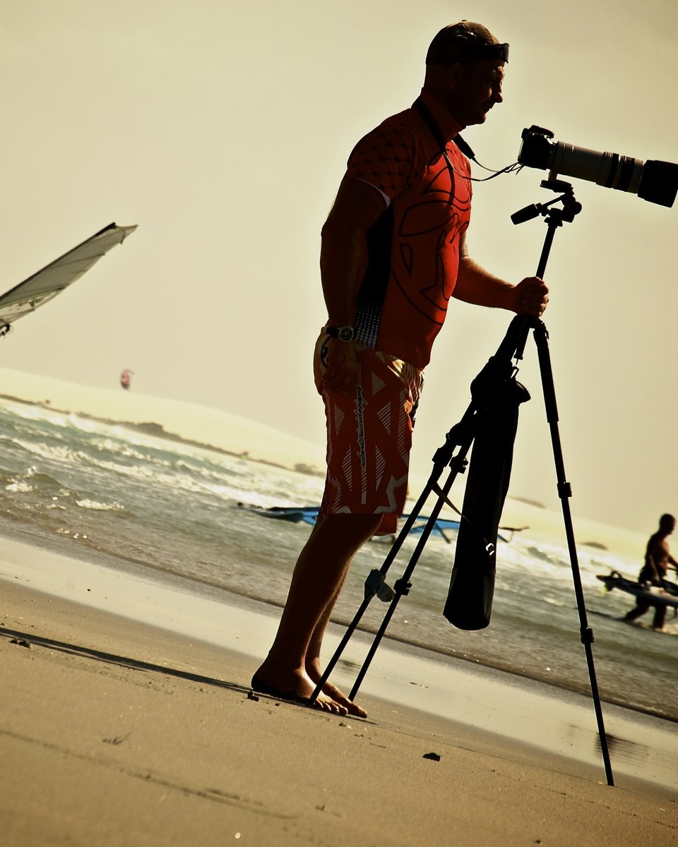 Fotógrafo com câmera no tripé filmando um windsurfista na praia de Jericoacoara ao entardecer.