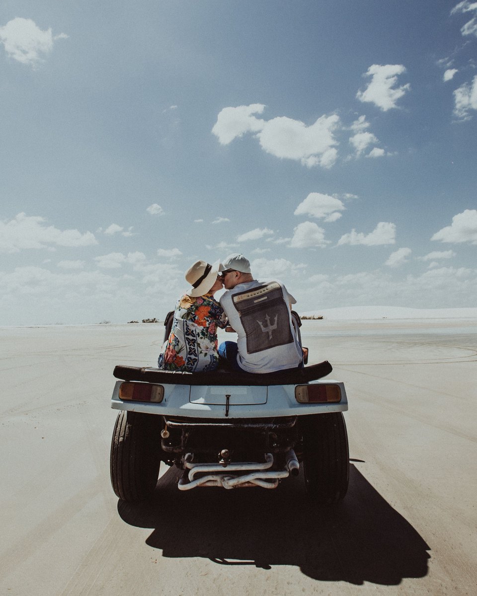 Casal de chapéu sentado abraçado na traseira de um buggy azul, atravessando a areia plana da praia sob céu azul com nuvens.