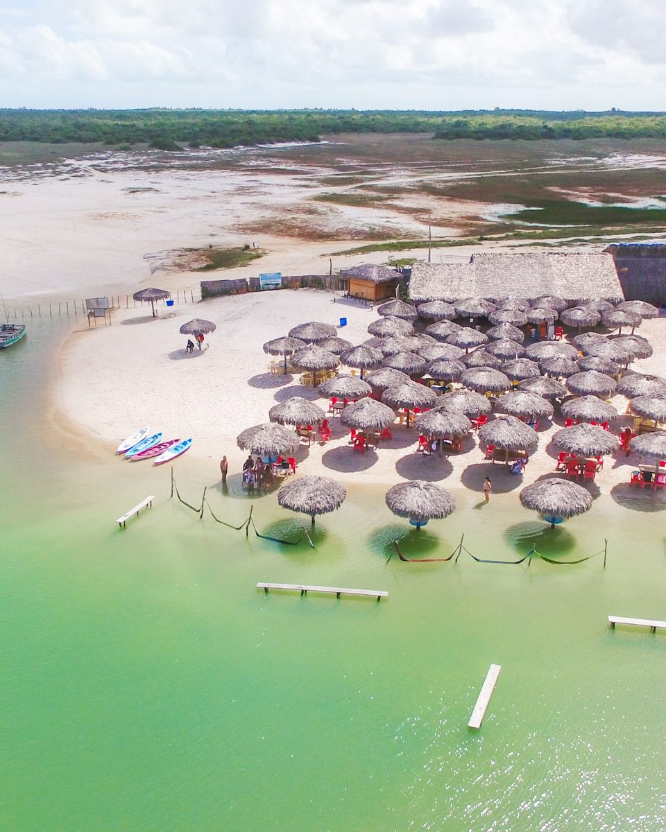 Vista aérea da Lagoa Azul, com a água verde-clara, dezenas de guarda-sóis de palha na margem de areia branca, um restaurante de telhado de palha e buggys estacionados.